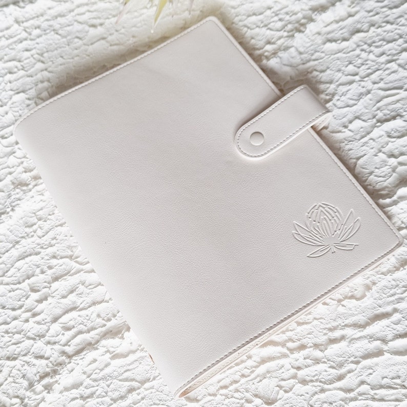 May include: A white leather planner with a floral design embossed on the front cover. The planner has a flap closure with a button.