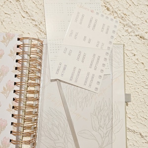 May include: A close-up of a planner with a floral design and a set of monthly calendar stickers. The stickers are labeled with the months of the year and are arranged in a vertical column.