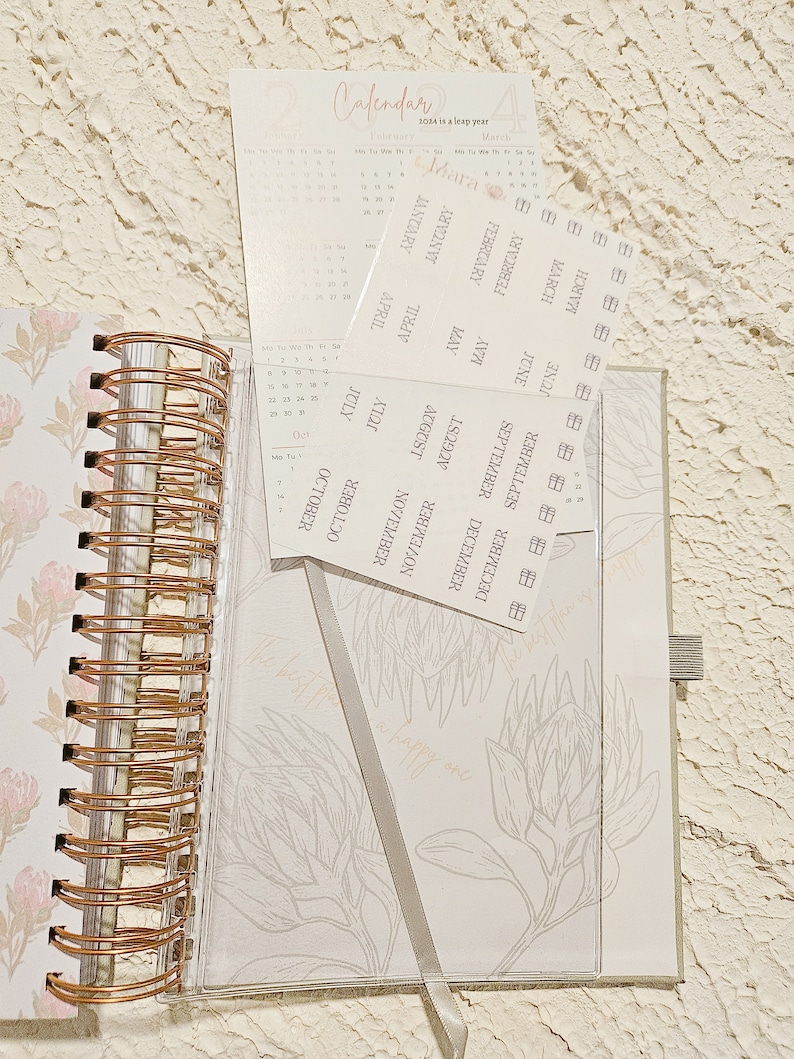May include: A close-up of a planner with a floral design and a set of monthly calendar stickers. The stickers are labeled with the months of the year and are arranged in a vertical column.