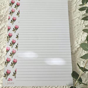 May include: A lined notepad with a floral border of pink and green flowers. The notepad has a white background with horizontal lines for writing. The bottom left corner has the text "Mara".
