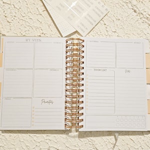 May include: A white weekly planner with a rose gold spiral binding. The planner has sections for each day of the week, a to-do list, and a notes section. The planner also has a section for priorities.