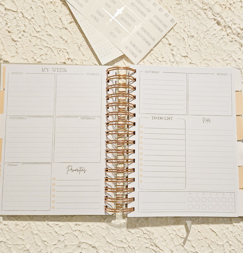 May include: A white weekly planner with a rose gold spiral binding. The planner has sections for each day of the week, a to-do list, and a notes section. The planner also has a section for priorities.