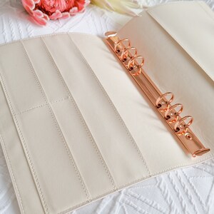 May include: A cream-colored leather bound planner with a rose gold ring binder. The planner is open and has several pockets and slots for holding papers and cards.