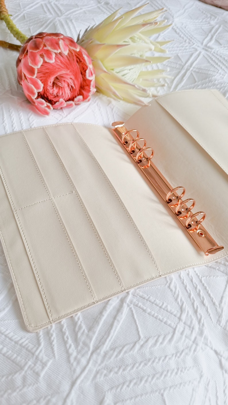 May include: A cream-colored leather bound planner with a rose gold ring binder. The planner is open and has several pockets and slots for holding papers and cards.