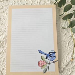 May include: A lined notepad with a cream border features a blue bird illustration perched on a branch with a pink flower. The notepad has a white background with horizontal lines for writing.