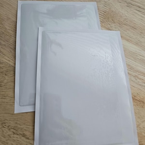 May include: Two white, rectangular, plastic sleeves with a smooth, glossy finish. The sleeves are designed to protect and display items.