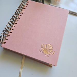 May include: A pink spiral bound notebook with a gold embossed flower design. The notebook has a white ribbon bookmark.