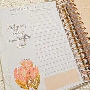 May include: A white planner with a gold spiral binding. The planner has a quote about next year's words awaiting another voice by T.S. Eliot. The planner has a pink floral illustration.