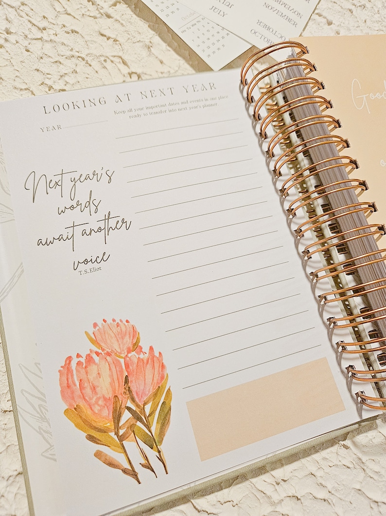 May include: A white planner with a gold spiral binding. The planner has a quote about next year's words awaiting another voice by T.S. Eliot. The planner has a pink floral illustration.