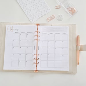 May include: A white monthly planner with a rose gold binding and the word "January" written in cursive at the top. The planner is open to the month of January and has a grid layout with days of the week labeled at the top.