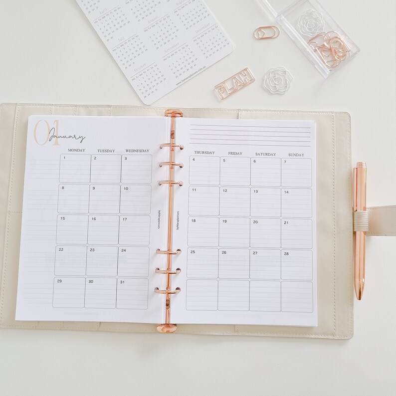 May include: A white monthly planner with a rose gold binding and the word "January" written in cursive at the top. The planner is open to the month of January and has a grid layout with days of the week labeled at the top.