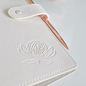 May include: White leather journal with a gold pen and a white embossed flower design.
