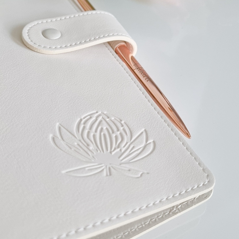 May include: White leather journal with a gold pen and a white embossed flower design.