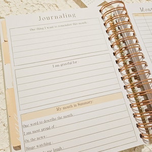 May include: A white journal with a gold spiral binding. The journal has a section for journaling with prompts such as "One thing I want to remember this month" and "I am grateful for".