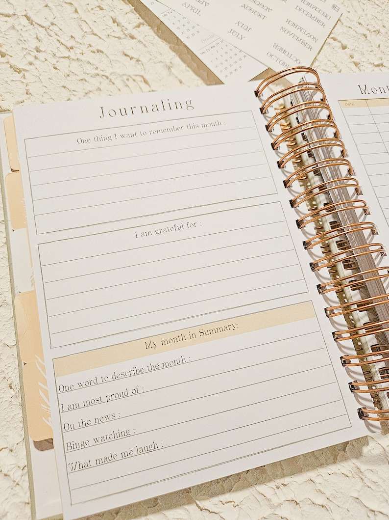 May include: A white journal with a gold spiral binding. The journal has a section for journaling with prompts such as "One thing I want to remember this month" and "I am grateful for".