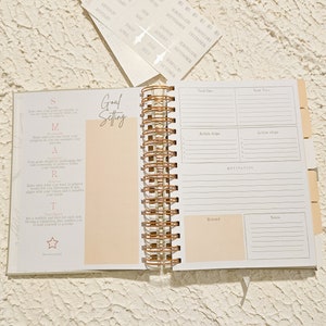 May include: A white and gold spiral bound journal with a peach colored page open to a goal setting section. The page has a list of steps to help set goals. The next page is a goal setting spread with sections for goal one, goal two, action steps, motivation, reward, and notes.