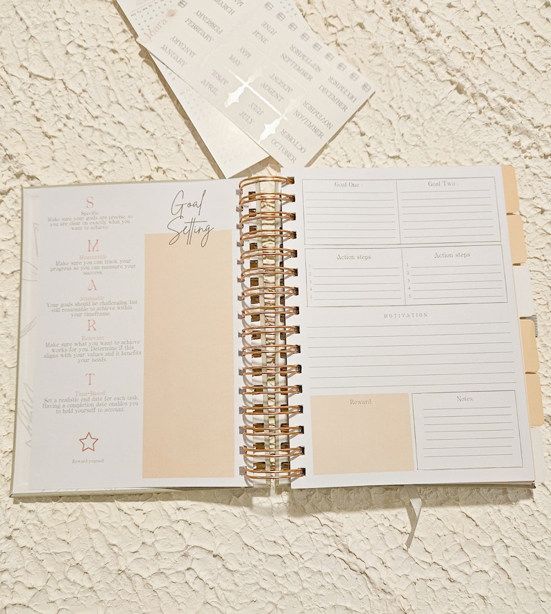 May include: A white and gold spiral bound journal with a peach colored page open to a goal setting section. The page has a list of steps to help set goals. The next page is a goal setting spread with sections for goal one, goal two, action steps, motivation, reward, and notes.