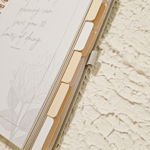 May include: A white planner with a gold spiral binding and gold tabs. The planner has a quote on the front that reads "An hour of planning can save you 10 hours of doing."