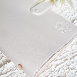 May include: White leather bound journal with a rose gold closure and a floral design embossed on the front cover.