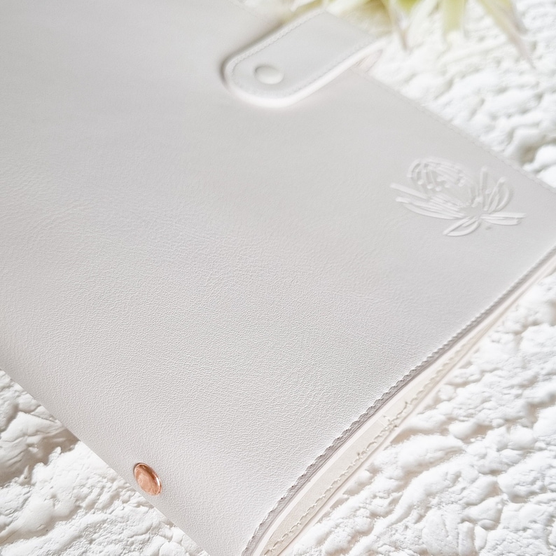 May include: White leather bound journal with a rose gold closure and a floral design embossed on the front cover.