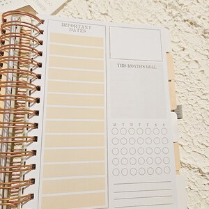 May include: A white planner with a copper spiral binding. The planner has sections for important dates, a monthly goal, and a calendar grid with days of the week labeled. The planner has a cream colored background.