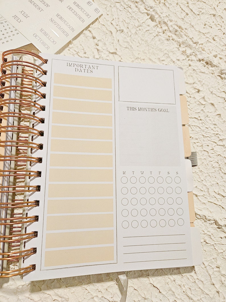 May include: A white planner with a copper spiral binding. The planner has sections for important dates, a monthly goal, and a calendar grid with days of the week labeled. The planner has a cream colored background.