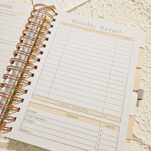 May include: A white and gold monthly budget planner with a spiral binding. The planner has sections for income, expenses, savings, and balance. The planner also has a section for a summary of expenses.