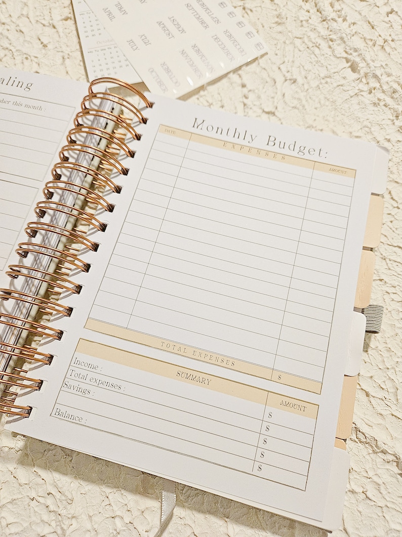 May include: A white and gold monthly budget planner with a spiral binding. The planner has sections for income, expenses, savings, and balance. The planner also has a section for a summary of expenses.