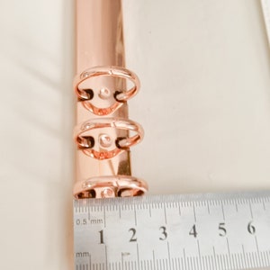 May include: Close-up of a rose gold binder ring mechanism with three rings. A ruler is placed below the rings, showing the measurement of 3 cm.