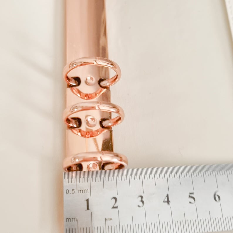 May include: Close-up of a rose gold binder ring mechanism with three rings. A ruler is placed below the rings, showing the measurement of 3 cm.