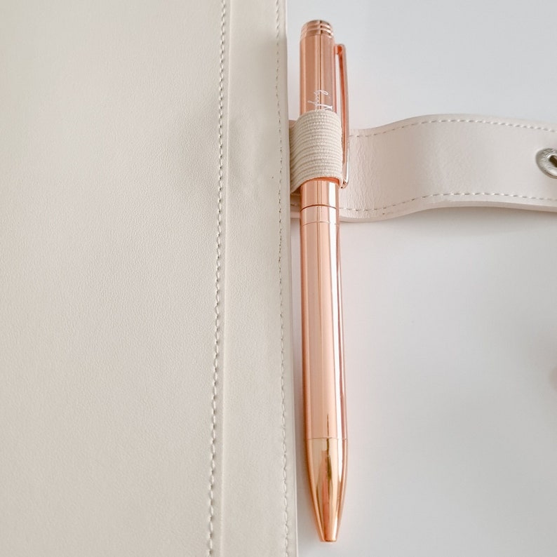 May include: A rose gold pen is held in place by a leather strap on a cream-colored leather journal.