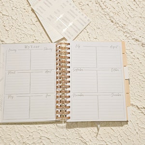 May include: An open white planner with a gold spiral binding. The planner has a monthly calendar layout with spaces for each month of the year. The months are labeled January through December.