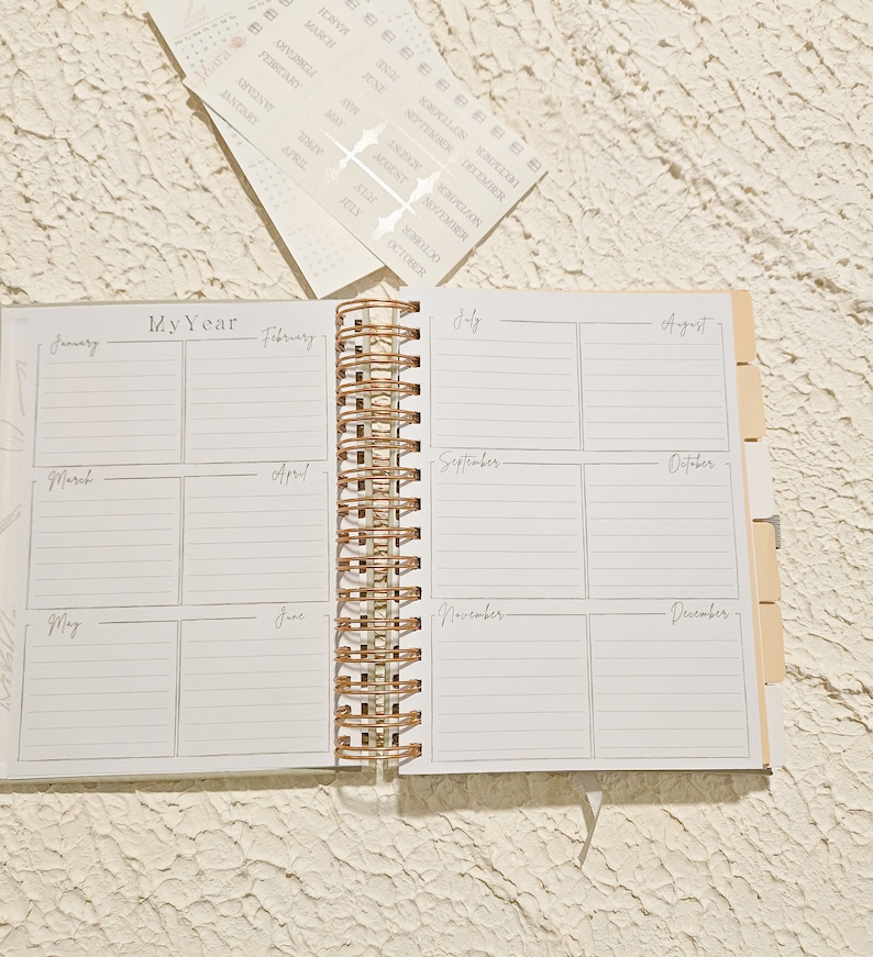 May include: An open white planner with a gold spiral binding. The planner has a monthly calendar layout with spaces for each month of the year. The months are labeled January through December.