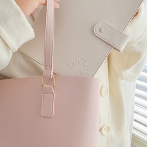 May include: A light pink tote bag with a gold clasp and a matching light pink strap. A cream-colored leather notebook with a floral design is tucked into the bag.