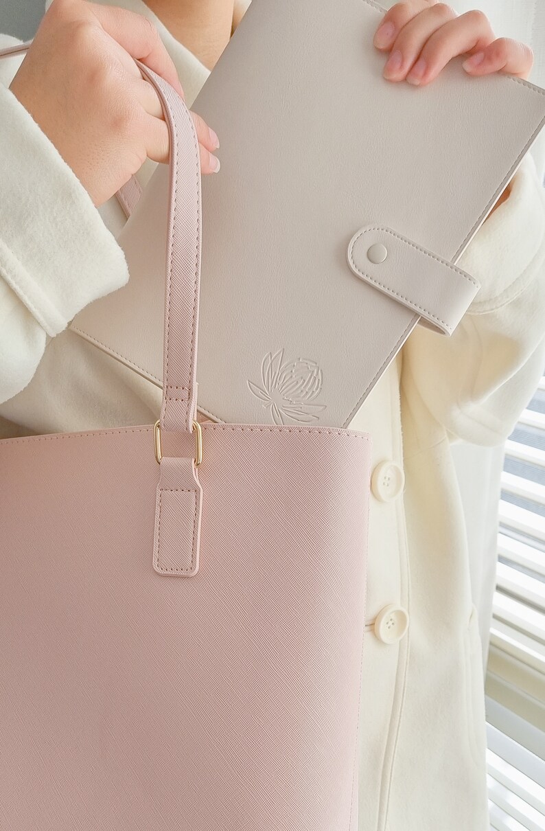 May include: A light pink tote bag with a gold clasp and a matching light pink strap. A cream-colored leather notebook with a floral design is tucked into the bag.