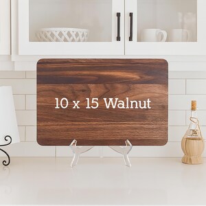 May include: A rectangular walnut cutting board, measuring 10 x 15 inches, is displayed on a clear acrylic stand. The board has a rich, dark brown wood grain. White text on the board reads "10 x 15 Walnut".