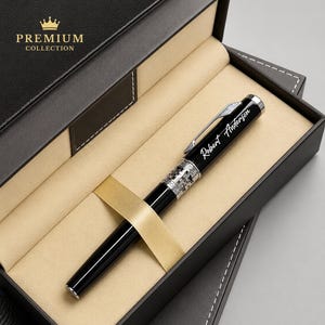 Puede incluir: Un bol&iacute;grafo negro con detalles plateados y el nombre "Robert Anderson" grabado, presentado en una caja de regalo negra con una cinta dorada. La caja est&aacute; etiquetada como "Premium Collection" con una corona dorada.