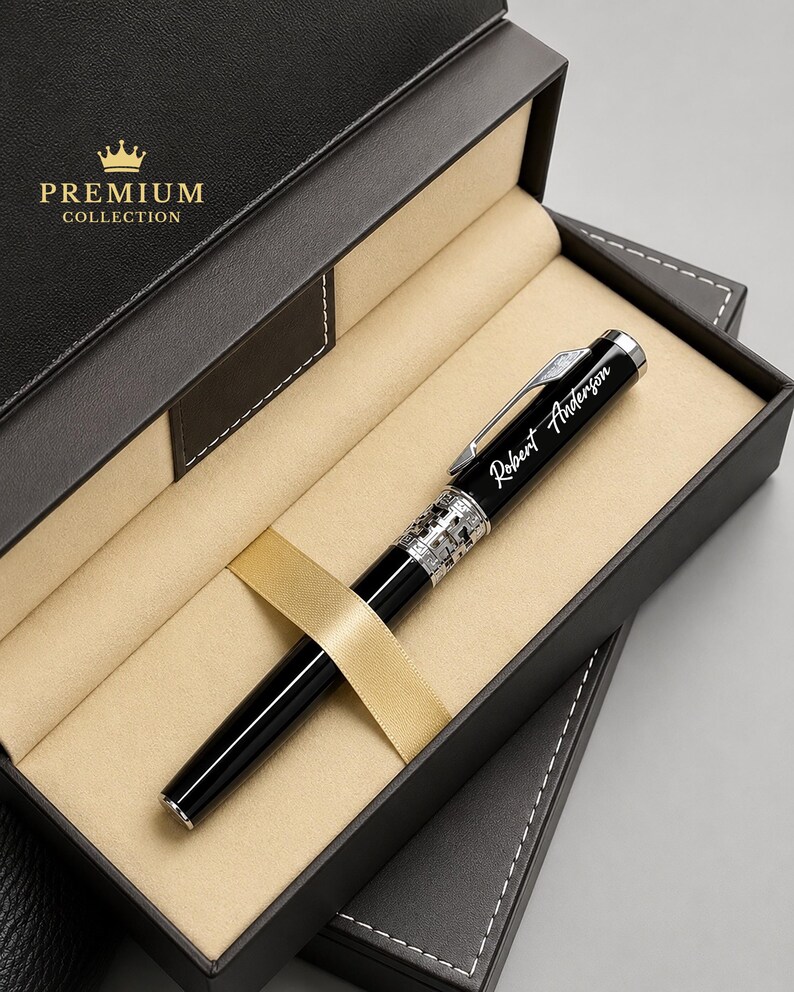 Puede incluir: Un bol&iacute;grafo negro con detalles plateados y el nombre "Robert Anderson" grabado, presentado en una caja de regalo negra con una cinta dorada. La caja est&aacute; etiquetada como "Premium Collection" con una corona dorada.
