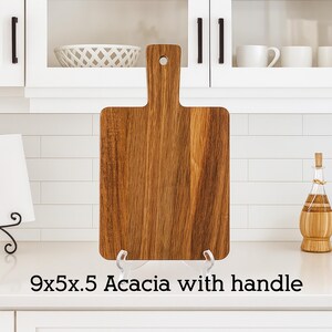 May include: A rectangular acacia wood cutting board with a handle. The board is 9 inches long, 5 inches wide, and 0.5 inches thick. The wood grain is visible. The text "9x5x.5 Acacia with handle" is at the bottom.