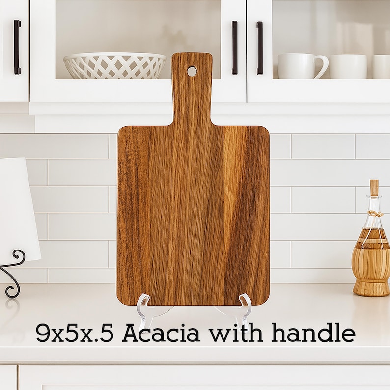 May include: A rectangular acacia wood cutting board with a handle. The board is 9 inches long, 5 inches wide, and 0.5 inches thick. The wood grain is visible. The text "9x5x.5 Acacia with handle" is at the bottom.