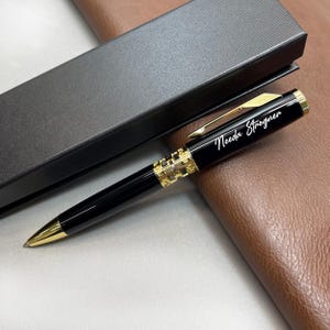 May include: Black and gold pen with the name "Needa Strogner" engraved on the barrel. The pen features a gold clip and accents, and a decorative gold band near the center. The pen is presented in a black gift box.
