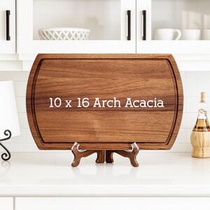 May include: A rectangular Arch Acacia cutting board, 10 x 16 inches, with a rounded edge and a juice groove. The board is displayed on a wooden stand, showcasing its rich brown wood grain. The text "10 x 16 Arch Acacia" is printed on the board.