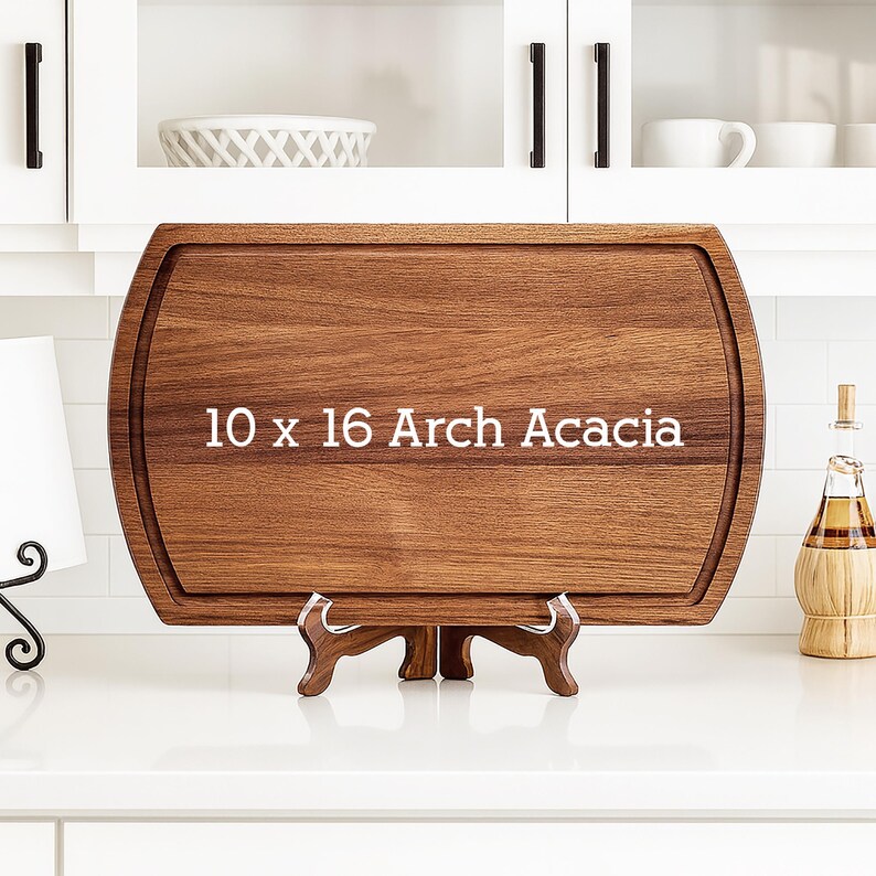 May include: A rectangular Arch Acacia cutting board, 10 x 16 inches, with a rounded edge and a juice groove. The board is displayed on a wooden stand, showcasing its rich brown wood grain. The text "10 x 16 Arch Acacia" is printed on the board.