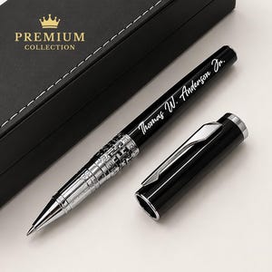 Puede incluir: Un bol&iacute;grafo negro con detalles plateados y el nombre grabado "Thomas W. Anderson Jr." se muestra junto a su tapa. El bol&iacute;grafo se presenta con una caja negra que tiene las palabras "PREMIUM COLLECTION" en letras doradas.