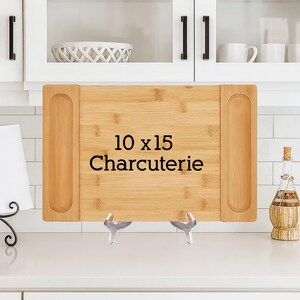 May include: A rectangular bamboo charcuterie board, 10 x 15 inches, with recessed handles. The words "10 x 15 Charcuterie" are printed in black. The board is displayed on clear stands against a white kitchen backdrop.