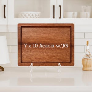 May include: A rectangular acacia wood cutting board, 7 x 10 inches, with a juice groove. The board is displayed on a clear stand against a white kitchen backdrop. The text "7 x 10 Acacia w/JG" is printed on the board.