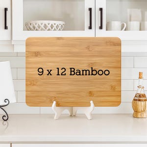 May include: A rectangular bamboo cutting board, 9 x 12 inches, with the text "9 x 12 Bamboo" in black. The board is displayed on a white stand. A decorative bottle with a cork stopper is on the right.
