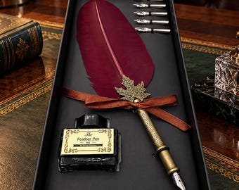May include: A burgundy feather pen set in a black box featuring a metallic maple leaf accent, a brown ribbon, five silver nibs, and a glass ink bottle with a label reading The Feather Pen Ink. The pen barrel is engraved with Elizabeth North.