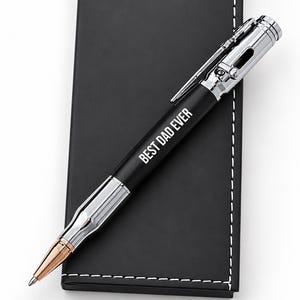May include: A black and silver ballpoint pen with a bullet-shaped design and a rose gold tip. The barrel features the white text BEST DAD EVER. The pen rests on a black rectangular case with white stitching and the text PREMIUM COLLECTION.