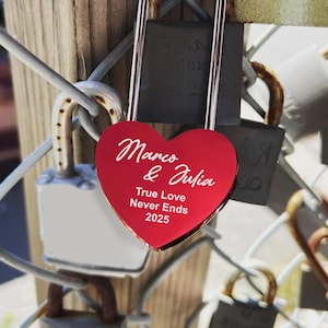 May include: A red heart-shaped padlock with the inscription "Marco & Julia True Love Never Ends 2025" attached to a metal fence with other padlocks.
